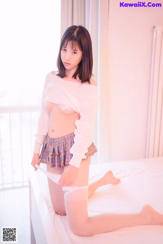 A woman in a white sweater and plaid skirt posing on a bed.
