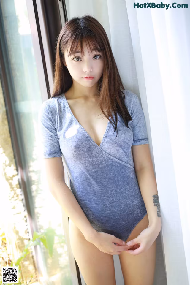 A woman in a gray shirt is posing by a window.